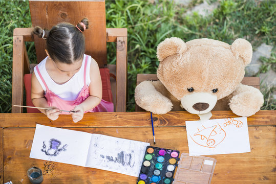 Adorable little girl painting outdoor in a sunny summer day with her teddy bear friend,  seen from above. Outdoor education concept
