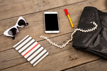 Naklejka premium women set with bag, smart phone, sunglasses, notepad, pen and pearl on brown wooden desk