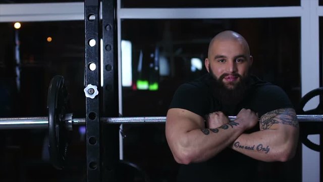Strong muscular bodybuilder resting on barbell in gym with happy smile on face