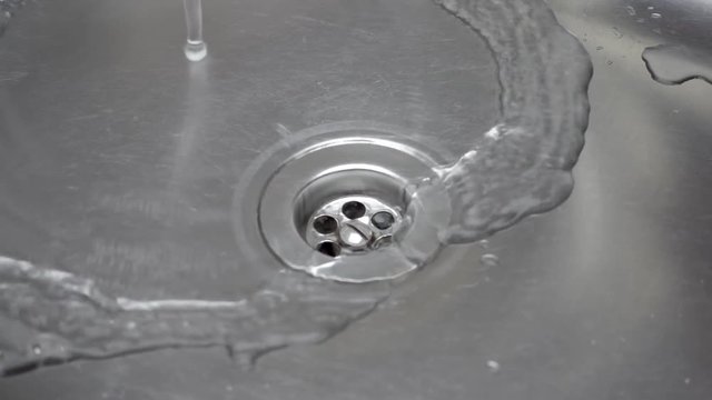 The stream of water falls from a tap in the sink in stainless steel and goes down the drain. Slow motion, high speed camera, 250fps