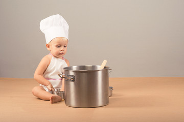 Charming and surprised kid in an apron and chef hat sitting next to a big pot of soup and holding something in his hand
