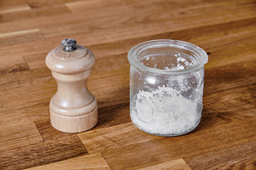 Pepper mill of light wood and a salt jar of transparent glass, standing on a table of dark brown laminated hardwood