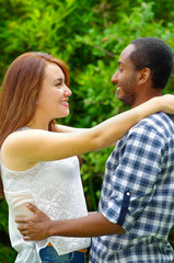 Interracial charming couple wearing casual clothes embracing and posing for camera in outdoors environment