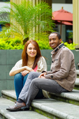 Interracial happy charming couple sitting on steps in front of building interacting and smiling for camera