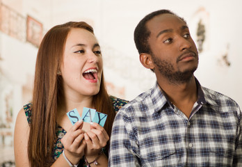 Charming interracial couple holding up small letters spelling the say while interacting happily, blurry studio background