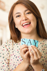 Headshot charming brunette woman holding up small letters spelling the word say and smiling to camera
