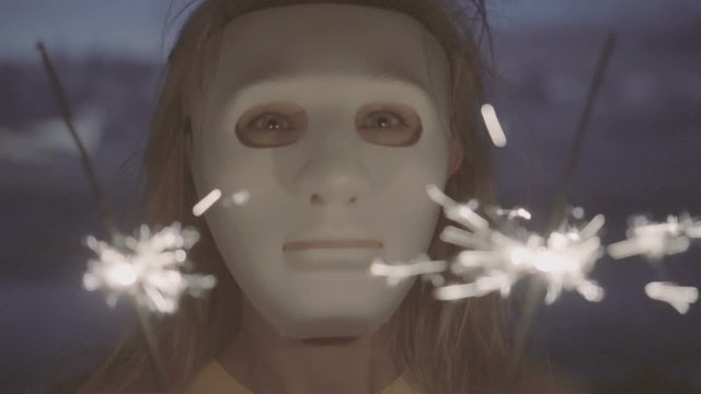 Closeup of scarry blonde woman wearing white mask with sparkler fireworks on the beach at twilight.