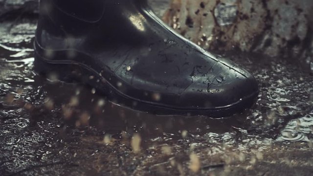 Black rubber boots step on a puddle, sunlit, and creates a splash which flies around. Slow motion, high speed camera, 250fps
