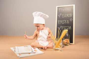 A little boy in a cap and apron cook cookbook studies. Beside him stand menu, spaghetti and colorful pasta