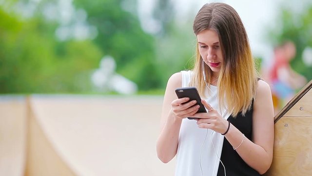 Young caucasian woman sending message and listen music outdoor at european city. Beautiful girl in sunglasses sitting on wooden bench using smartphone