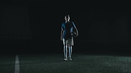 Young soccer player holding a ball and walking towards the camera then standing and looking straight in the dark stadium