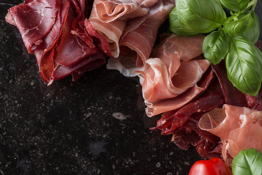 Jambon mix. Ham. Traditional Italian and Spanish salting, smoking, dry-cured dish - jamon Serrano and prosciutto crudo sliced with herbs and tomatoes on dark stone background. Copy space. Closeup. 
