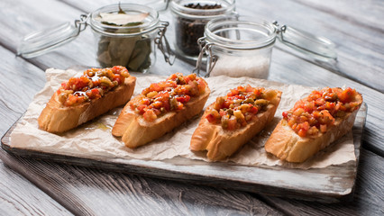 Pieces of baguette on paper. Jars with seasonings. Bruschetta with baked vegetables. Tasty meal on wooden table.