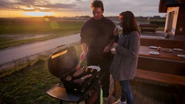 A couple is cooking on a barbecue grill. There is a beautiful sunset in the background. A woman offers a bit of meat to her husband. They're enjoying their time together.
