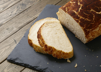 A loaf and slices of crusty white bread on a rustic slate chopping board and wooden table top background