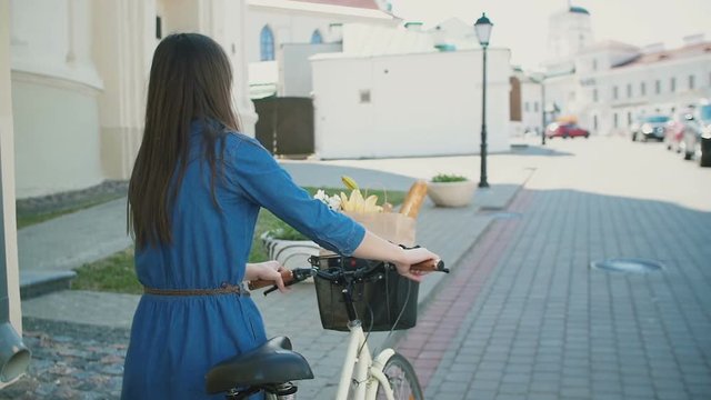 Back view of a girl with waving hair walking her bike with flowers and bread in a basket, slow mo, steadicam shot