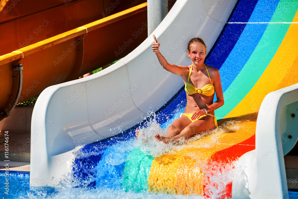 Wall mural Wet child on water slide at aquapark shows thumb up on water slides with flowing water in water park. Summer water park holiday. Outdoor.