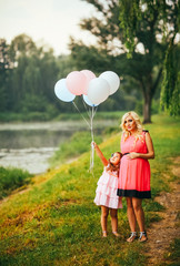 happy pregnant mother is walking with her little daughter in the park, happy family photo. Mother hold baloons. Family walk neaar lake. Beauty blond mother.