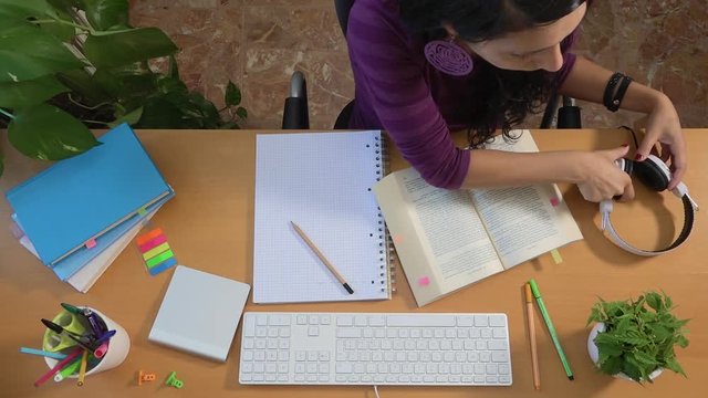 Female college student wearing headphones and studying to preparing exam. Caucasian girl listening lesson and writing on pad paper. Studying for degree. Young people, university life and learning