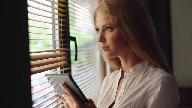 Close-up of charming blonde woman holding touch pad while standing near window in modern interior