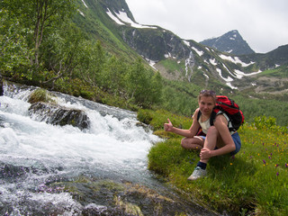 Woman in shorts and with backpack sitting on the bank of the creek and smiling on a background of mountains