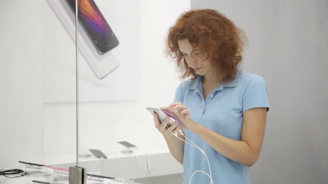 Young woman flipping a mobile phone in a digital technology shop, picture through the glass of the showcase