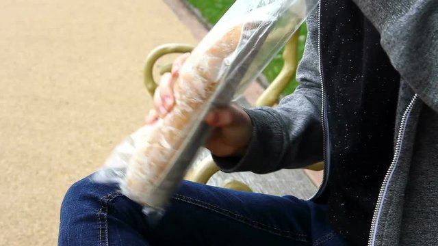 Young adult or late teens woman seated on a bench seat in a park, opening a shop bought roast chicken salad sub roll sandwich packet prior to eating it.