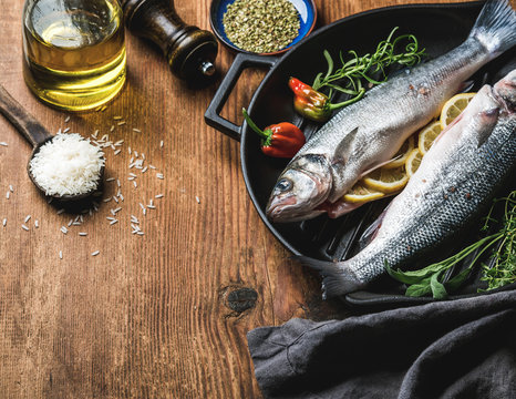 Ingredients for cookig healthy fish dinner. Raw uncooked seabass  with rice, olive oil, lemon slices, herbs and spices on black grilling iron pan over rustic wooden background, copy space