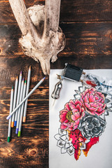 beautiful sketch drawing flowers with colored pencils lying on old wooden background, tattoo machine and skull of a goat