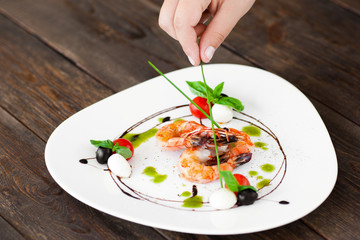 Hand decorated plate with grilled shrimps with green sticks, dark wooden background. Food preparation before serving at restaurant