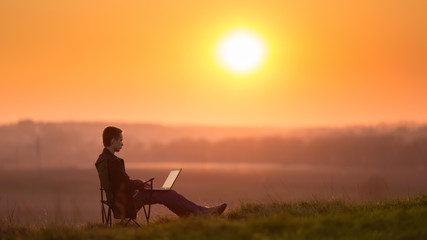 Fototapeta na wymiar The man sit and work on the laptop by sunset (sunrise) background on the top of the hill