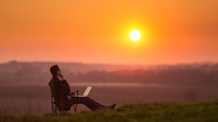 The man sit and work on the laptop by sunset (sunrise) background on the top of the hill