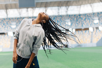 Young african american teenage girl in gray hoodie listening to music and playing with her hair at stadium - Powered by Adobe