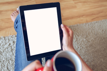 Woman in jeans sitting on the carpet near the sofa using a tablet, drinking coffee from a red cup. Online education concept. e-learning. back view