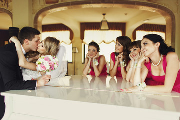 Groom bends bride over a piano and kisses her while bridesmaids