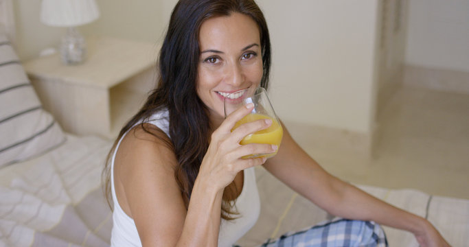 Smiling healthy young woman drinking a glass of freshly squeezed orange juice as she sits on her bed in the morning