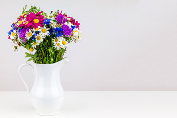 Wildflowers in white ceramic jug with copy space. Wild flower bouquet on white table. Bunch of wild herbs and flowers in a white jug. Wild flowers in a vase.