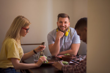 Happy friends spending their free time in vegan restaurant or cafe. Smiling man looking at camera while his friends eating healthy vegan dishes.