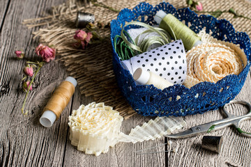  Sewing.   Colored ribbons and spools of thread in blue openwork basket on sackcloth and on gray wooden table.