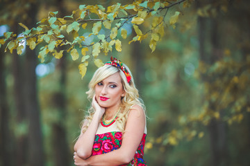 Girl in red dress standing on the background of trees in the forest. Woman in red dres and bandana. Dress painted flowers. Model in summer meadow forest\park. Advertising photoshoot.