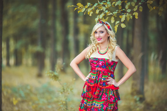 Girl in red dress standing on the background of trees in the forest. Woman in red dres and bandana. Dress painted flowers. Model in summer meadow forest\park. Advertising photoshoot.