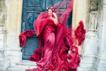 Beautiful young woman in a bright red dress and a crown of white spring flowers posing against the backdrop of the ancient door. Wind fanning,overblow, fan cloud dress. Brunet girl  with wrath. 