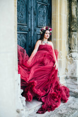 Beautiful young woman in a bright red dress and a crown of white spring flowers posing against the backdrop of the ancient door. Wind fanning,overblow, fan cloud dress. Brunet girl  with wrath. 