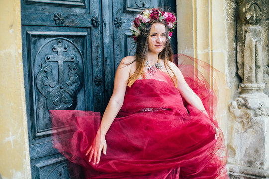 Beautiful young woman in a bright red dress and a crown of white spring flowers posing against the backdrop of the ancient door. Wind fanning,overblow, fan cloud dress. Brunet girl  with wrath. 