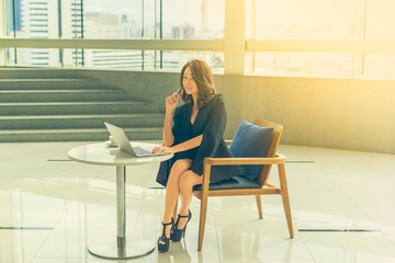 Beautiful Young Asian Businesswomen using a laptop computer on a table with a cup of hot coffee and pen sitting on the chair with happiness and concentrate.Vintage tone with city view background.