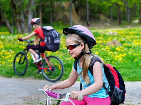 Children wearing bicycle helmet and glasses with rucksack rides bicycle. Bicyclist children is looking forwaqrd. Children ride on green grass and flowers in park outdoor.