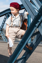 Little French School Child at Play / Retro styled cute boy at the way to - from school wear satchel, suspenders, shirt, shorts and red beret hat, play with paper airplane and climb at blue steel beam