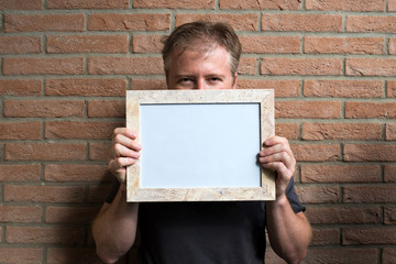 man holds a white wooden frame with blank space, brick wall background - free space for adding custom text or image
