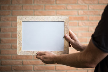 male hands holding a white wooden frame with blank space, brick wall background - free space for adding custom text or image