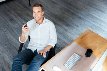 Cheerful young businessman sitting and using mobile phone at workplace
